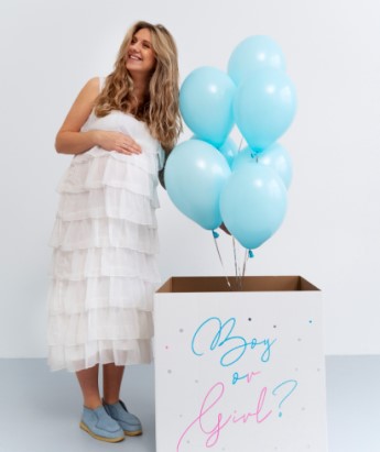 kobieta w ciąży w białej sukni otwiera box z napisem "boy or girl?" dla gender party i wylatują niebieskie balony